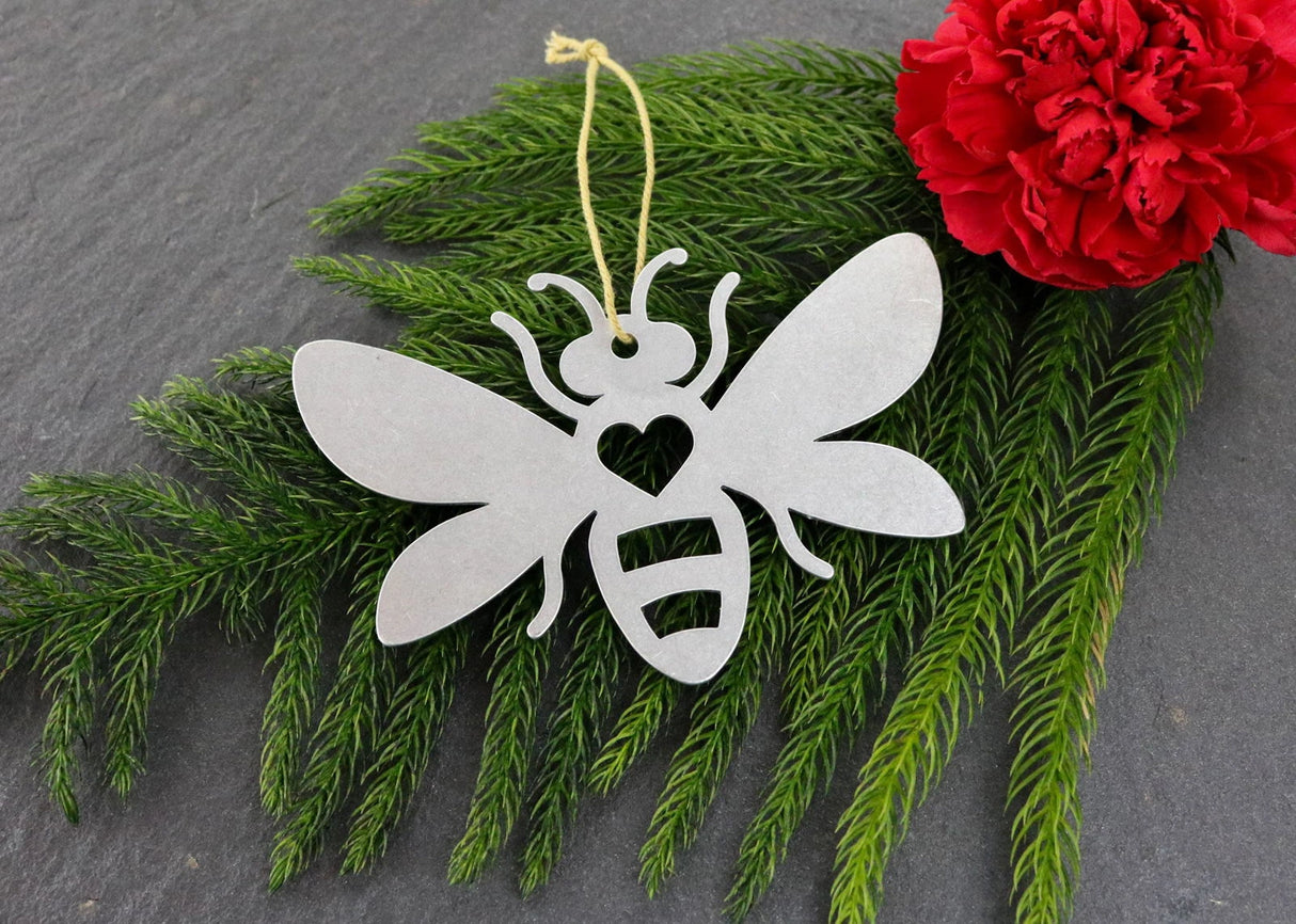 Bee metal ornament with heart cutout, surrounded by greenery and a red flower.