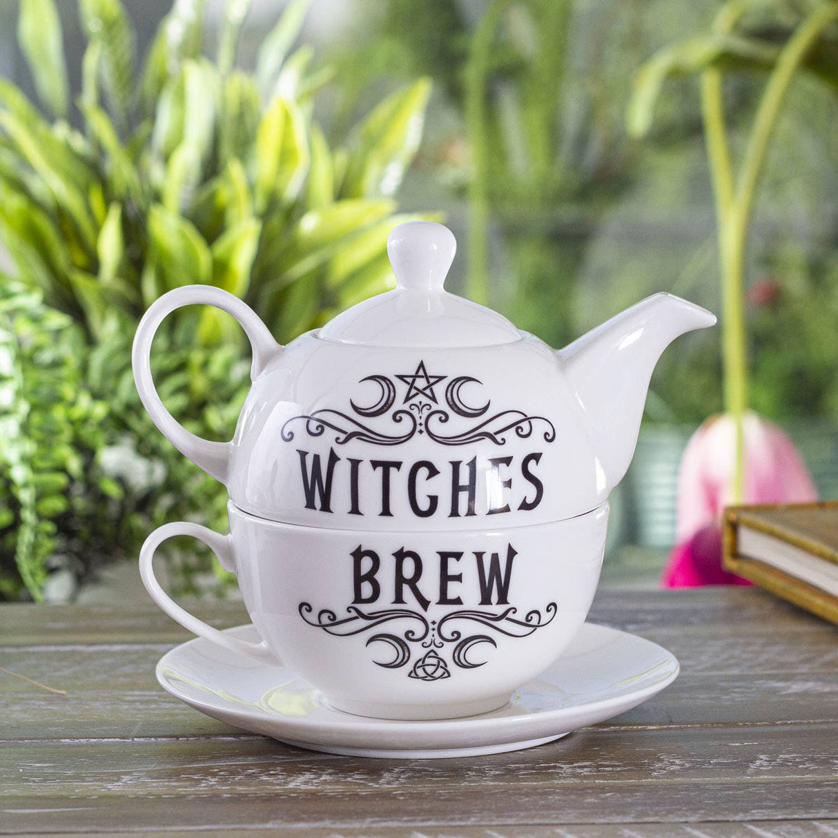 A white ceramic teapot and matching cup and saucer set stacked together on a wooden table, with the words 'WITCHES BREW' printed in black decorative lettering. The background shows a softly blurred garden setting with green foliage and a pink flower.