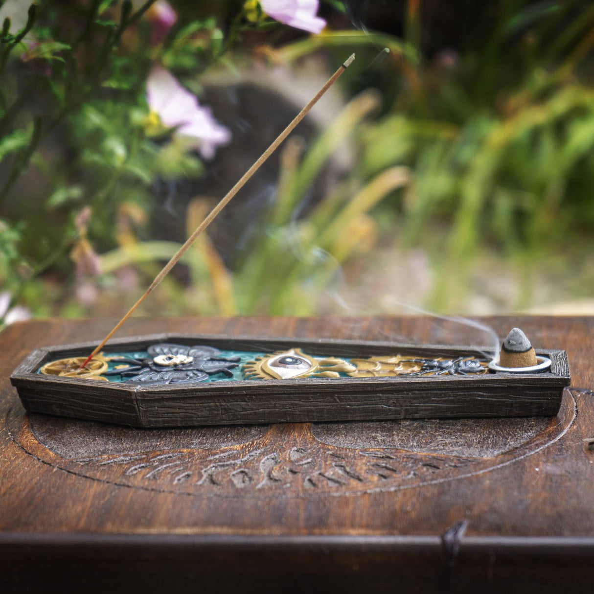 A decorative incense holder with a single burning incense stick, placed on a carved wooden surface with a blurred garden background.