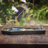 A decorative incense holder with a single burning incense stick, placed on a carved wooden surface with a blurred garden background.