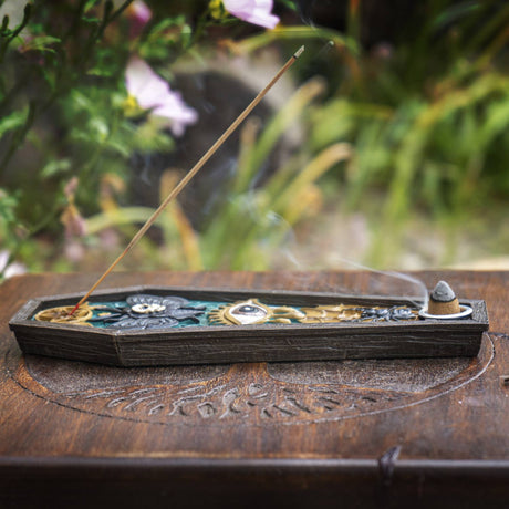 A decorative incense holder with a single burning incense stick, placed on a carved wooden surface with a blurred garden background.