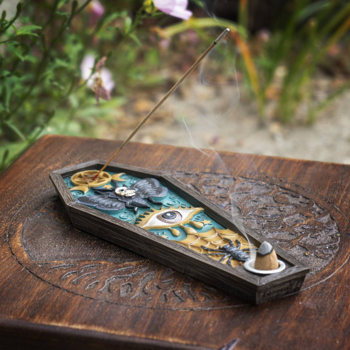 A coffin-shaped incense holder with intricate celestial and eye motifs, shown outdoors on a carved wooden surface with a burning incense stick resting inside.