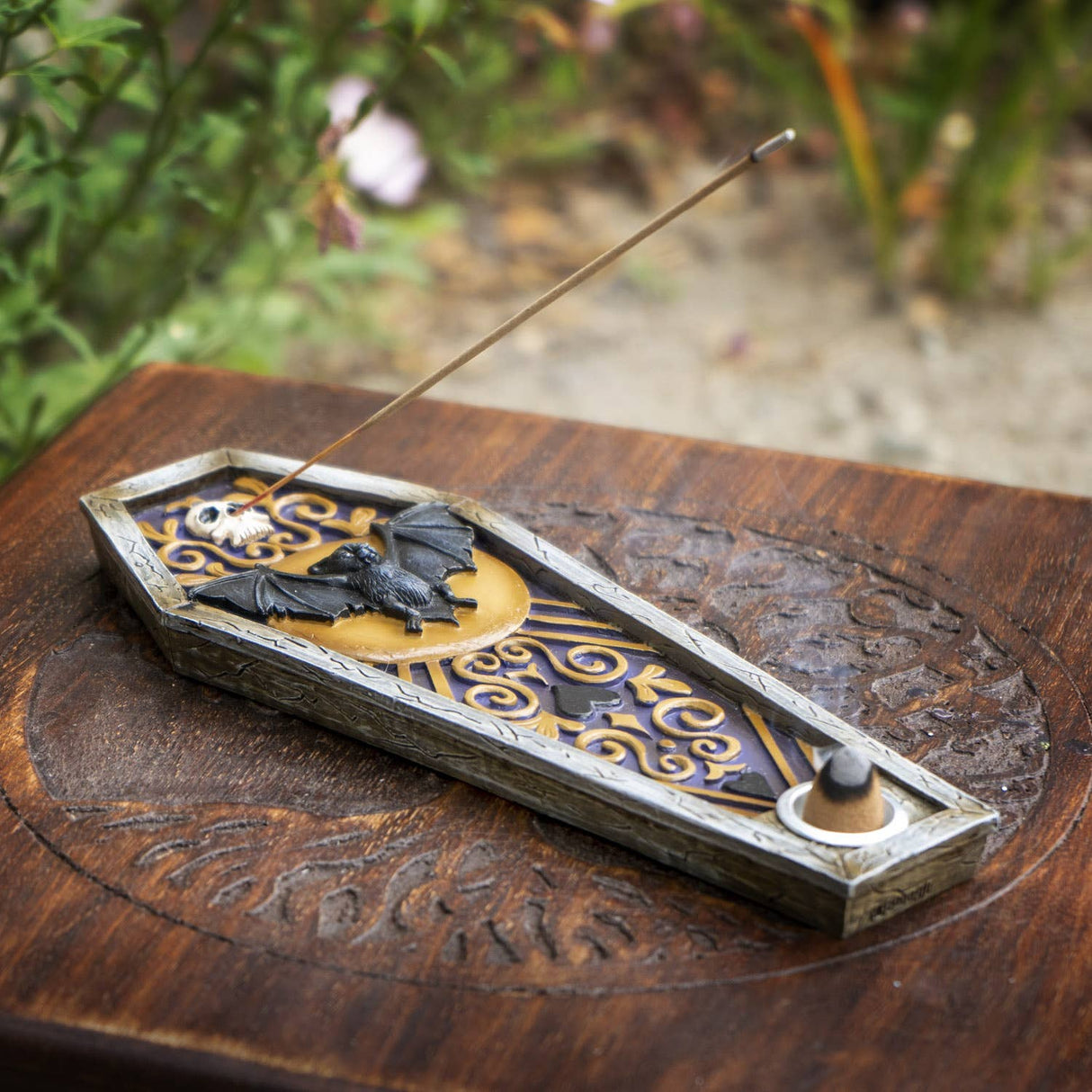 A coffin-shaped incense holder with bat and moon motifs, shown on a carved wooden surface with a blurred garden background. A incense stick rests across the top.