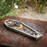 A coffin-shaped incense holder with bat and moon motifs, shown on a carved wooden surface with a blurred garden background. A incense stick rests across the top.