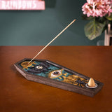 A coffin-shaped incense holder with gothic illustrations of a large eye, black spiders, and gold details, shown on a wooden table with an incense stick inserted.