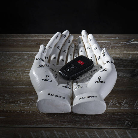 A pair of white sculptural hands with astrological and zodiac markings, holding a black car key fob on a rustic wooden surface against a dark background.
