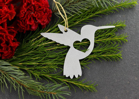 Hummingbird metal ornament with heart cutout, surrounded by greenery and red flowers.