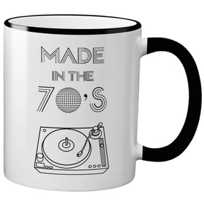 White ceramic mug with black text that reads "MADE IN THE 70'S" and a disco ball and record player graphic. The mug has a black rim and handle.