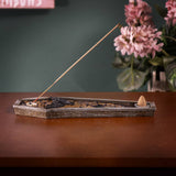 A coffin-shaped incense holder with a carved floral pattern, shown on a wooden table with a burning incense stick and a small cone incense holder inside. The background features a dark green wall and a blurred vase of pink flowers.
