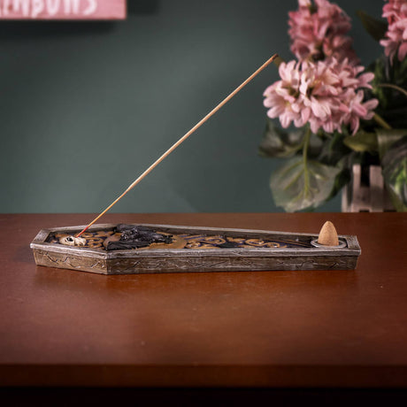 A coffin-shaped incense holder with a carved floral pattern, shown on a wooden table with a burning incense stick and a small cone incense holder inside. The background features a dark green wall and a blurred vase of pink flowers.