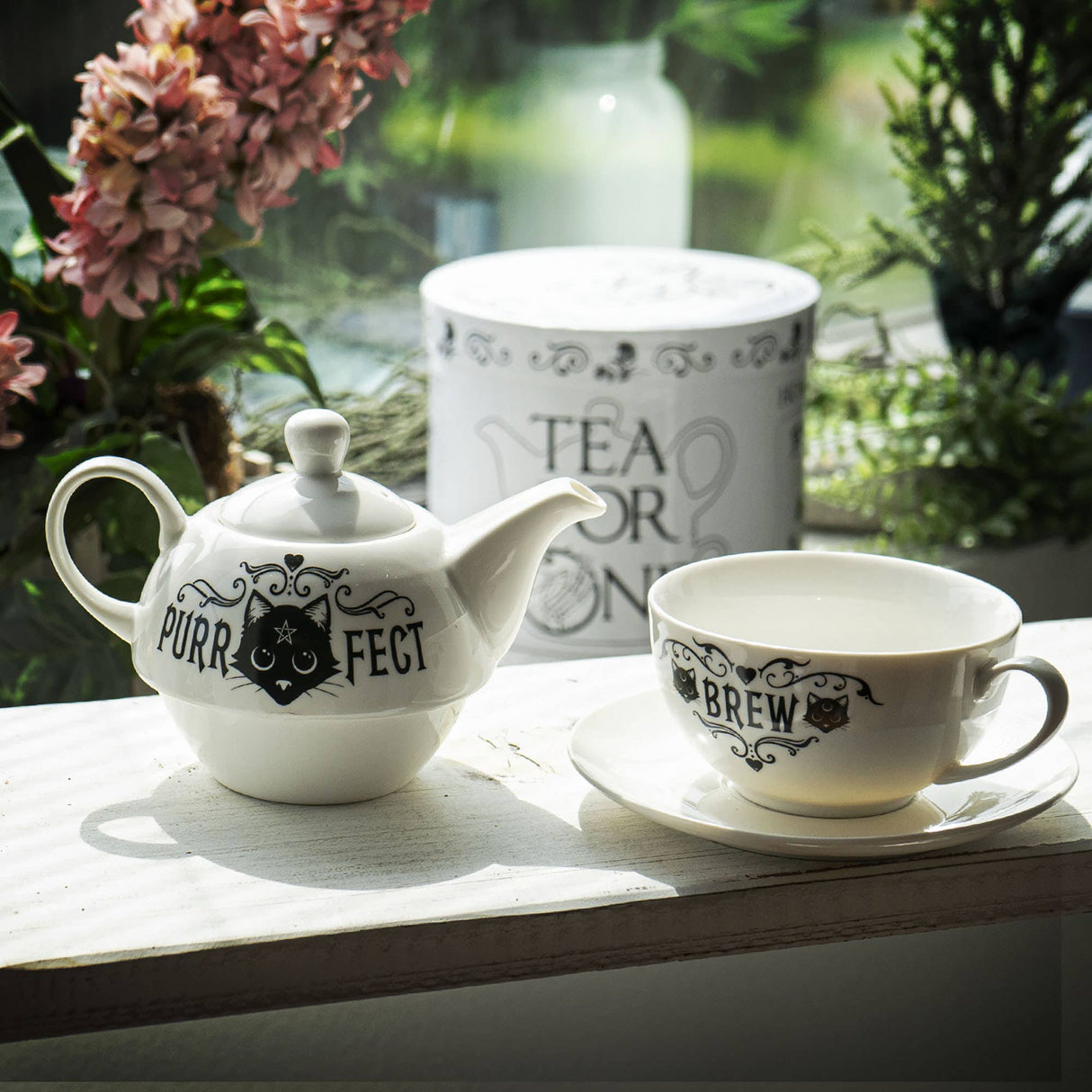 White teapot and cup with 'Purrfect' and 'Brew' text on a wooden surface with plants in the background.