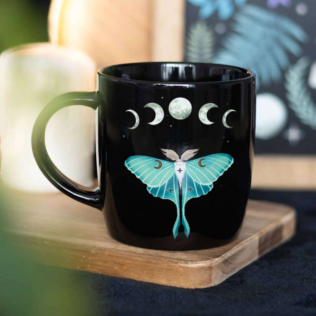 Black mug with moon phase design and butterfly illustration on a wooden coaster.