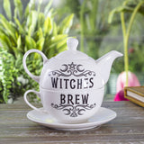 A white ceramic teapot and matching cup and saucer set stacked together on a wooden table, with the words 'WITCHES BREW' printed in black decorative lettering. The background shows a softly blurred garden setting with green foliage and a pink flower.