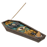 A coffin-shaped incense burner with a dark wood-look exterior and an open interior featuring a teal and gold mystical design with a large eye, black bat, spider, skull, and a small incense cone holder. A incense stick is inserted into the top opening, photographed on a plain white background.