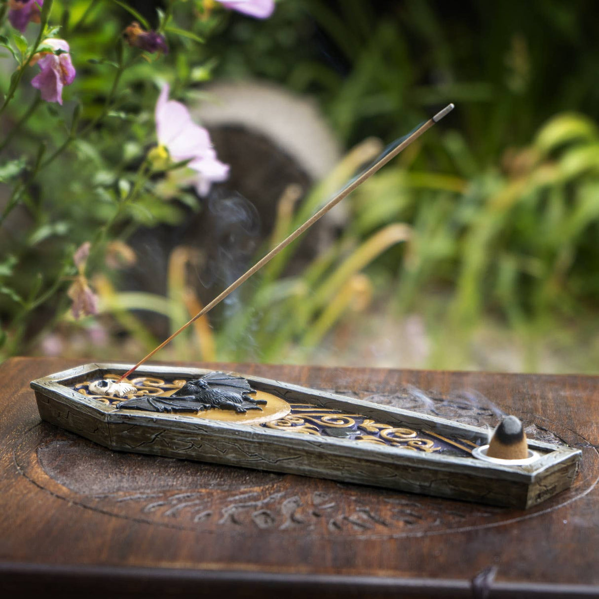 An ornate coffin-shaped incense holder with gold filigree details and a burning incense stick, set on a carved wooden table with a blurred garden background.